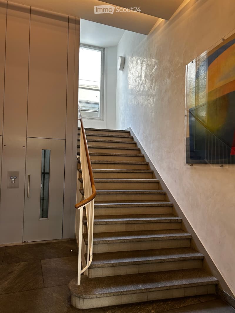 Modern staircase with beige stone steps, wooden handrail, and a wall-mounted light fixture. The hallway features a textured wall and a glass panel on one side.
