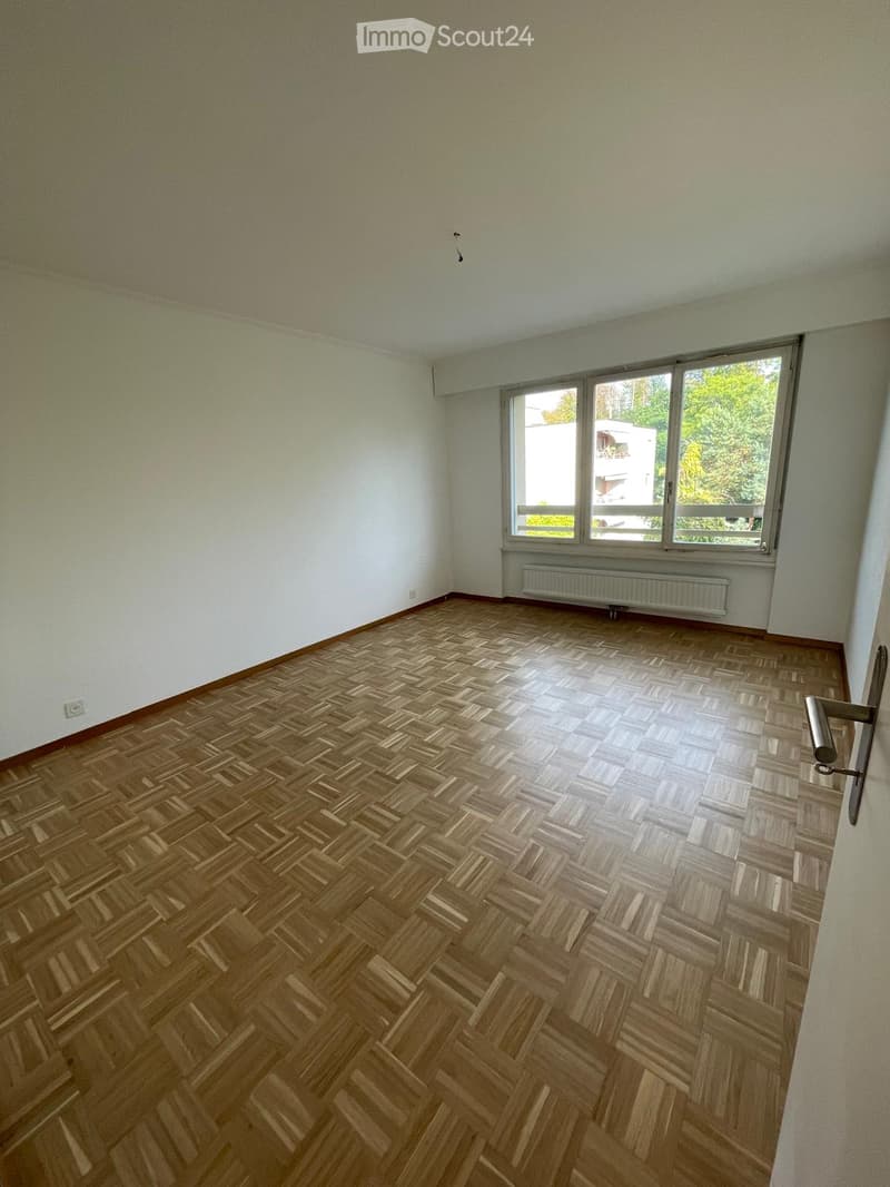 Empty room with wooden herringbone flooring, white walls, large window with view of greenery, radiator below window, and a door handle visible on the right side.