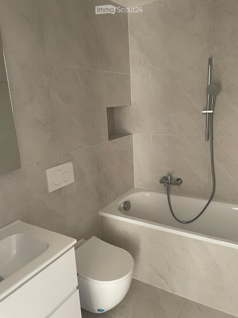 Modern bathroom with a bathtub, toilet, and showerhead. The walls are tiled in a light beige color, and the floor is also tiled in a matching shade. There is a small shelf built into the wall above the bathtub.