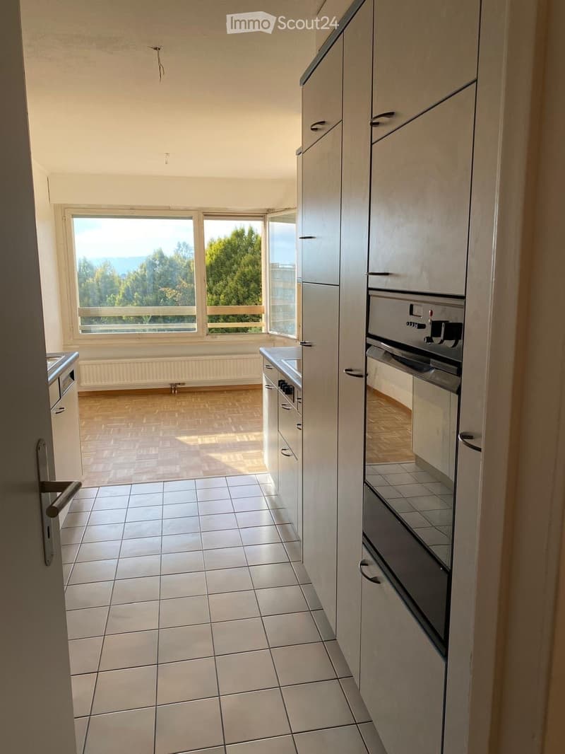 Empty kitchen with light wood cabinets, tiled floor, and large windows overlooking a scenic view.