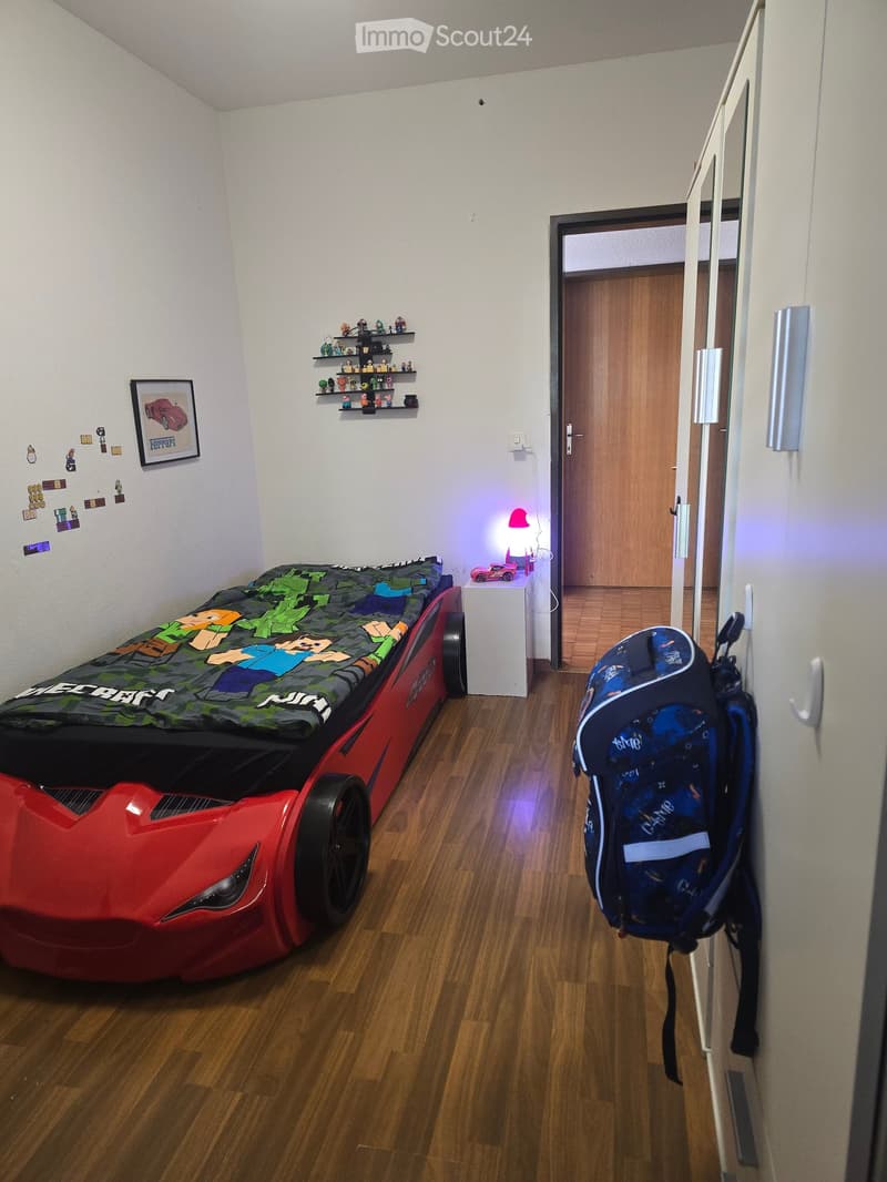 A child's bedroom with a car-shaped bed, a backpack hanging on the closet, a small shelf with toys, a lamp, and a picture frame on the wall.