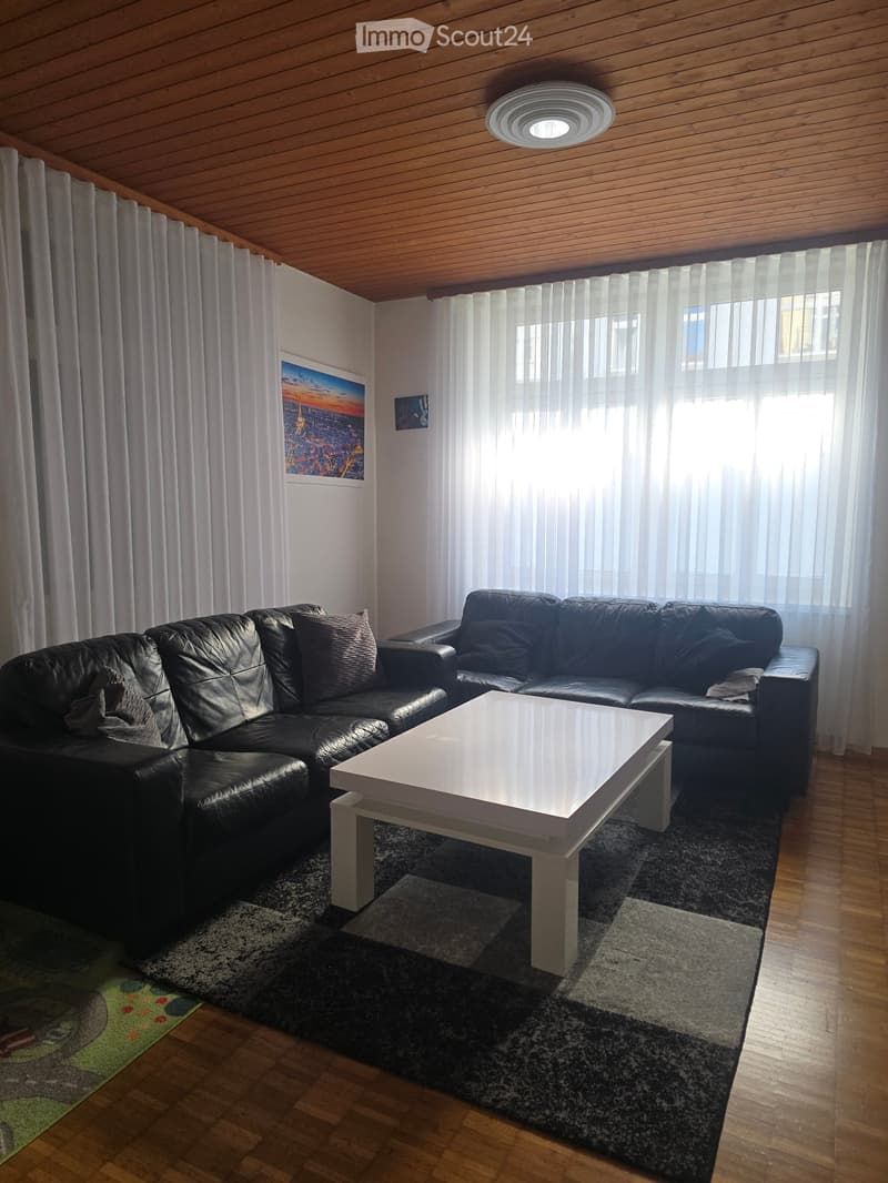 Modern living room with black leather sofa, white coffee table, and large windows with sheer curtains. Wooden floor and slanted wooden ceiling.