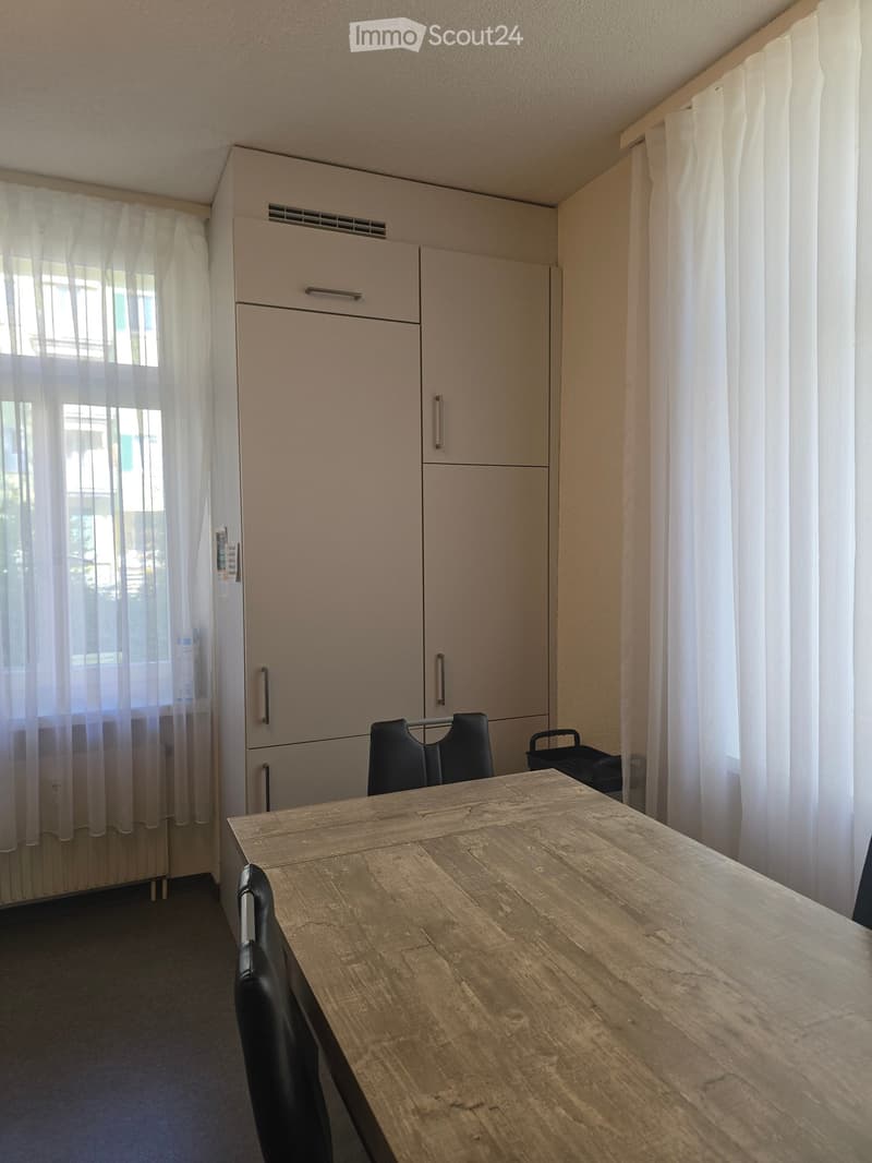 A small, furnished room with a wooden table and black chairs, a large wardrobe, and sheer curtains on the windows.