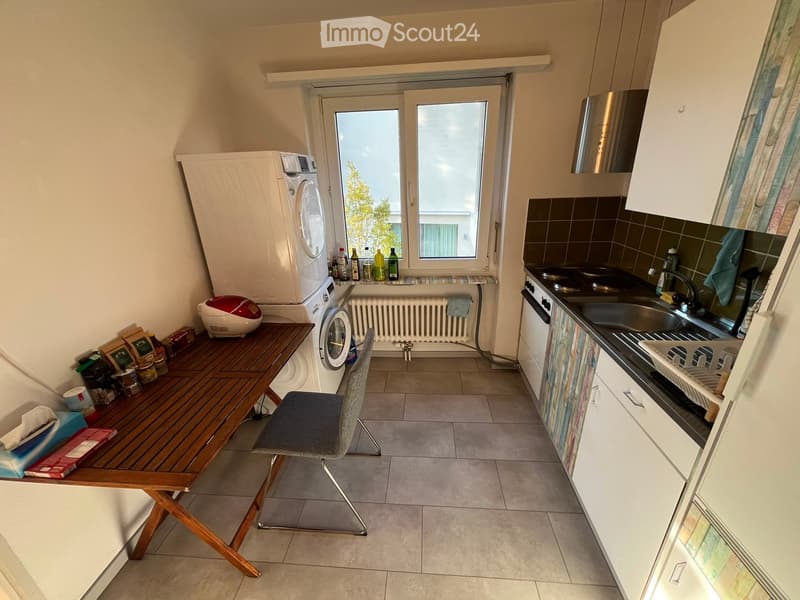 A compact kitchen with white cabinets, a wooden table, a chair, a washing machine, and a sink.