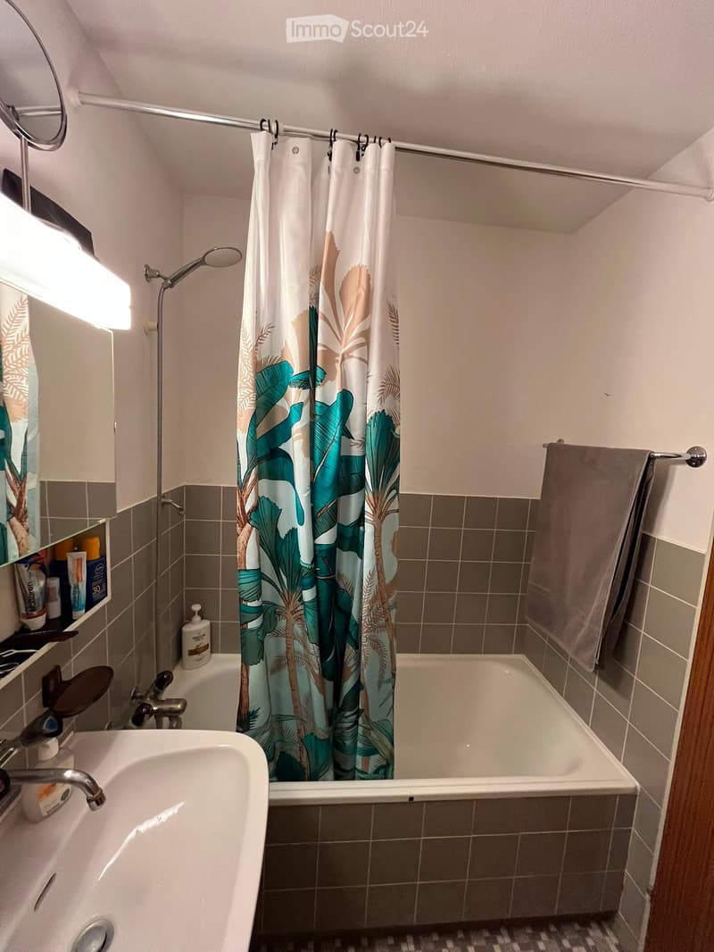 Bathroom with a bathtub, shower curtain, sink, mirror, and towel. The walls are tiled in a neutral color scheme, and the shower area features a modern design with a handheld showerhead.