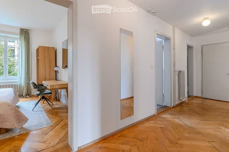 A furnished room with a bed, desk, chair, wardrobe, and mirror. The room features wooden flooring and white walls.