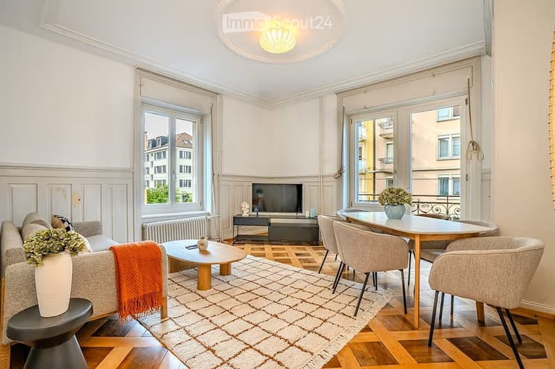 Bright living room with large windows, wooden flooring, a sofa with a throw, coffee table, dining table with chairs, and a television set.