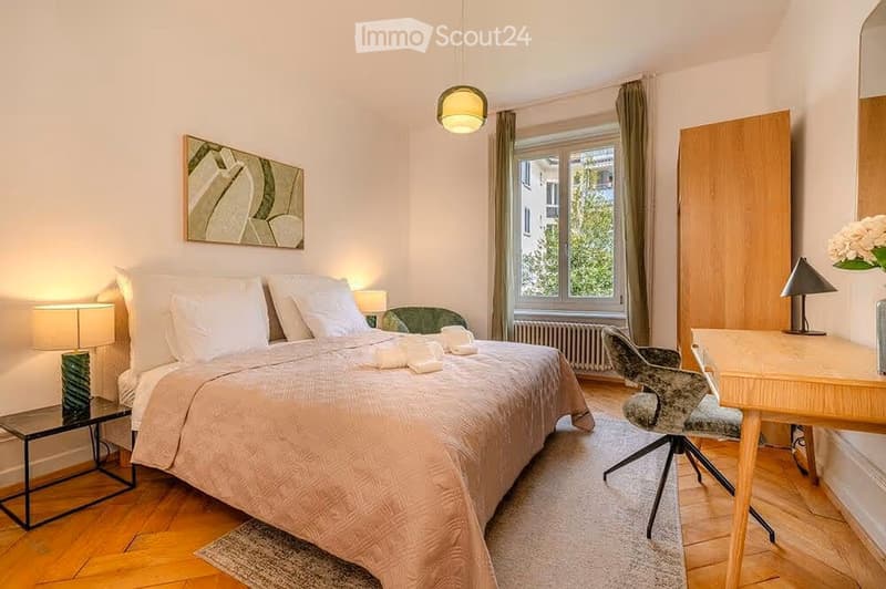 Cozy bedroom with a large bed, nightstand, desk, chair, and a window with curtains. The room features wooden flooring, a hanging light fixture, and a decorative picture on the wall.