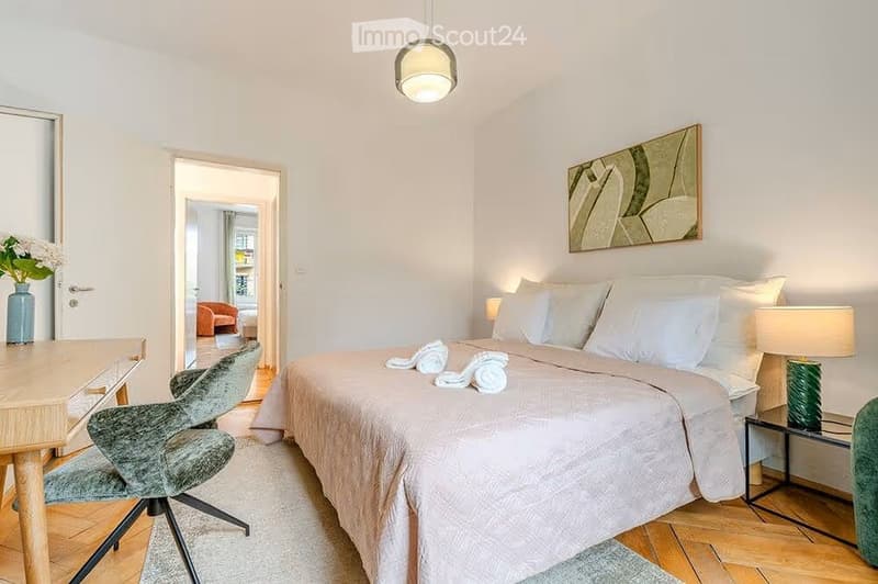 Cozy bedroom with a large bed, nightstand, chair, desk, and a view into an adjacent living area.