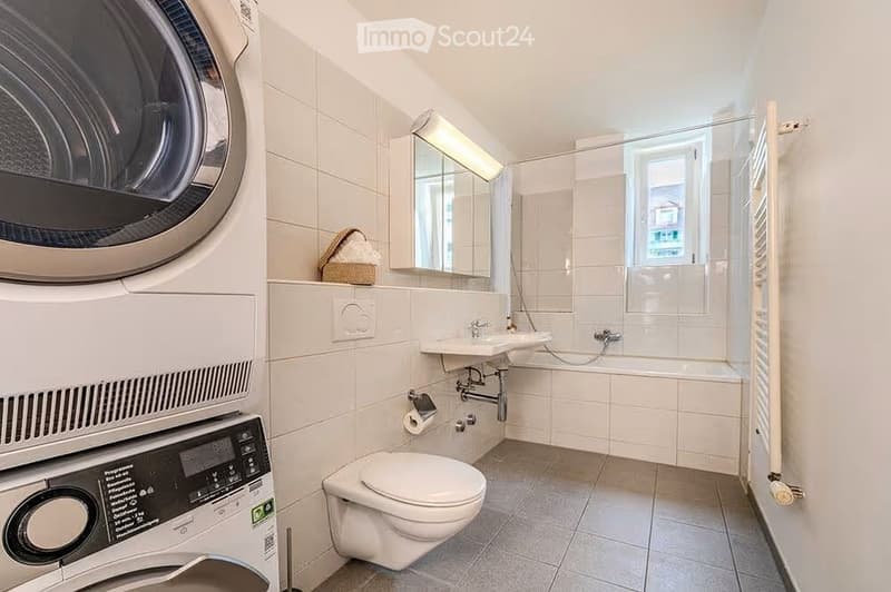 Modern bathroom with white tiles, a toilet, sink, mirror, washing machine, and a shower area with a window.
