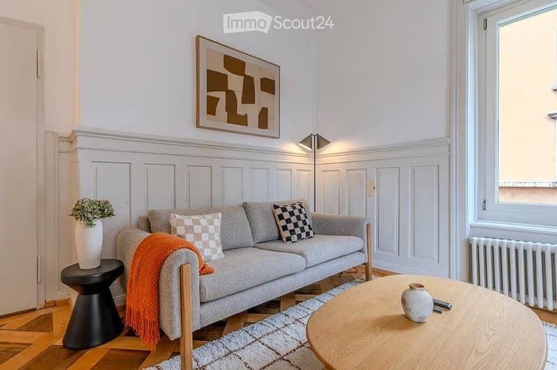 Modern living room with grey sofa, wooden coffee table, black stool, decorative vases, and artwork on the wall.