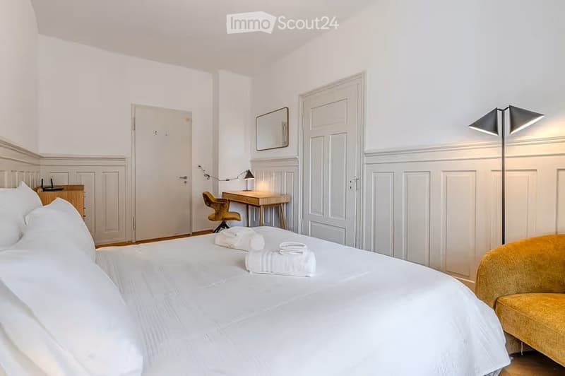 Elegant bedroom with a large white bed, neatly arranged towels, a yellow armchair, a wooden desk with a chair, a mirror, and a floor lamp.