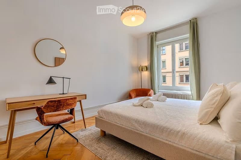 Cozy bedroom with a wooden bed, desk, chair, and a round mirror on the wall. The room features a large window with curtains and a modern pendant light.
