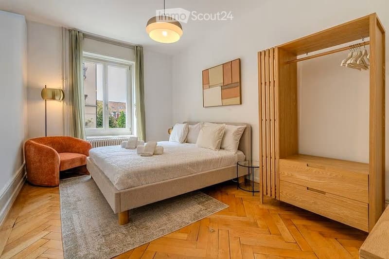 Cozy bedroom with a wooden bed, armchair, wardrobe, and large window with view.