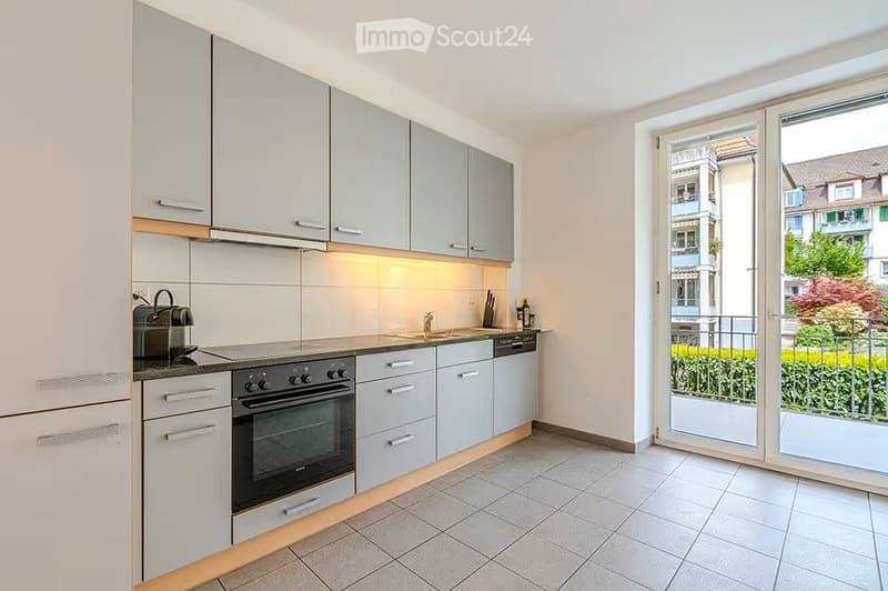 Modern kitchen with light grey cabinets, stainless steel appliances, and a large window opening to a balcony.