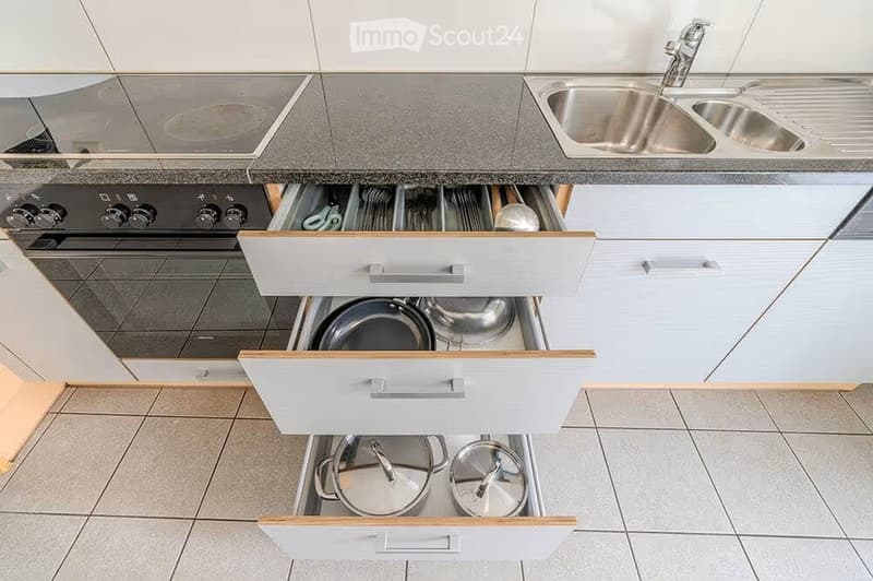 Modern kitchen with white cabinets, stainless steel sink, and open drawers revealing neatly organized pots and pans.