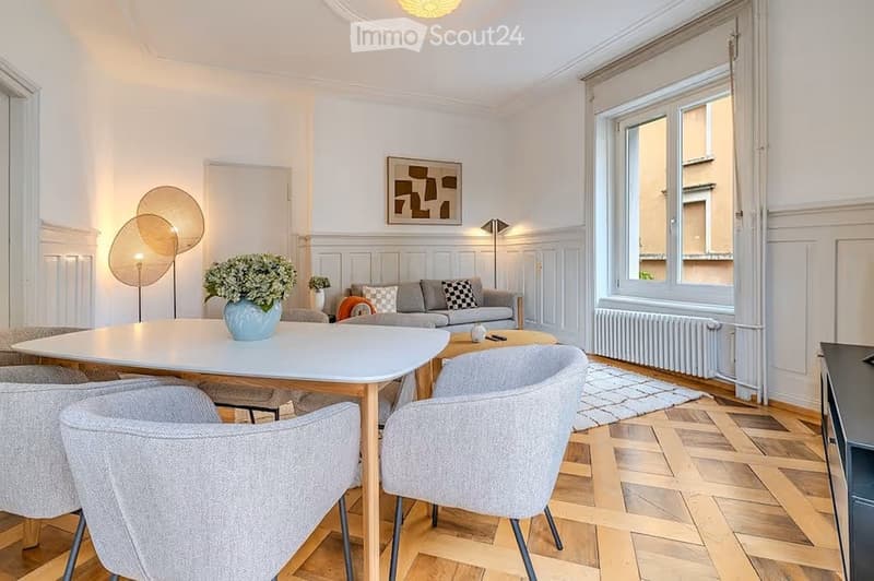Modern living room with light-colored walls, wooden flooring, and large windows. Features a sofa, coffee table, dining table with chairs, decorative vase, floor lamp, and artwork on the wall.