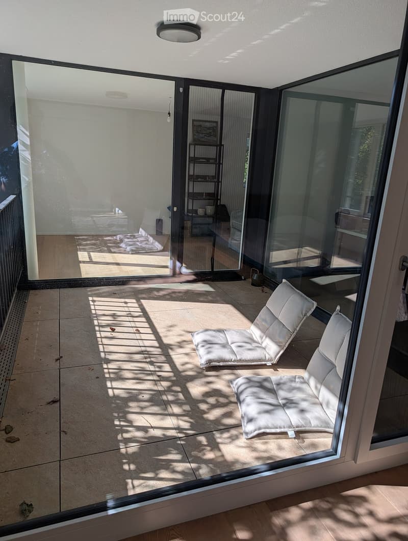 Modern balcony with tiled floor, large glass sliding doors, two white cushioned chairs, and a small shelf visible through the glass.