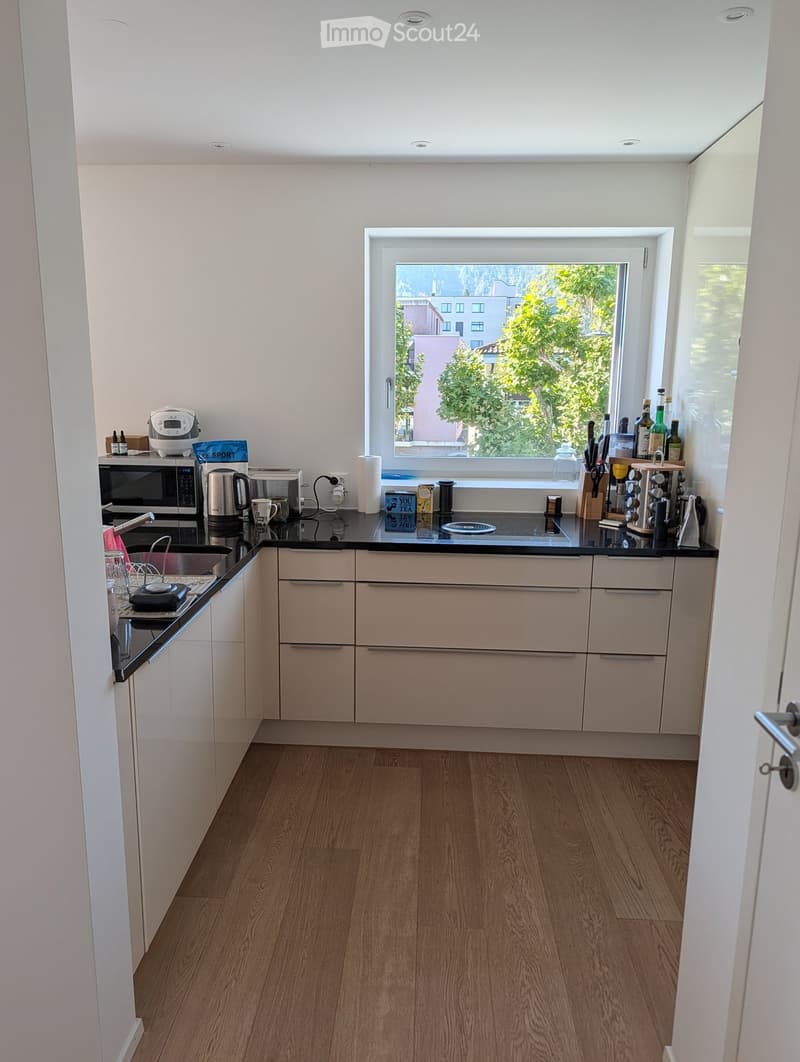 Modern kitchen with light-colored cabinets, wooden flooring, and various appliances including a microwave, toaster, coffee maker, and knife block. A window offers a view of greenery outside.