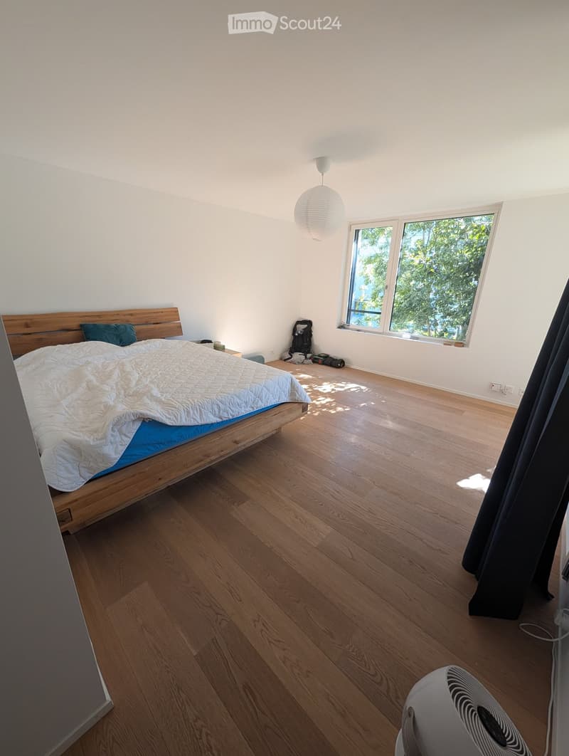 Bright bedroom with wooden flooring, large window, bed with white quilt and blue pillow, nightstand, fan, and backpack near the window.