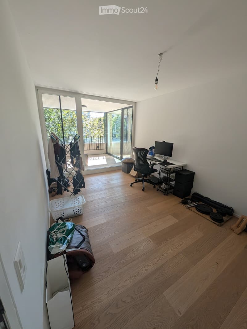A well-lit room with wooden flooring, a desk with a computer, a chair, a clothing rack, a laundry basket, and gym equipment in the corner.