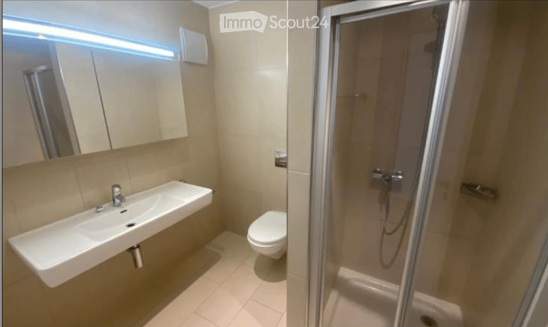 Modern bathroom with a white sink, toilet, mirror, and a glass-enclosed shower. The walls and floor are tiled in a light beige color.