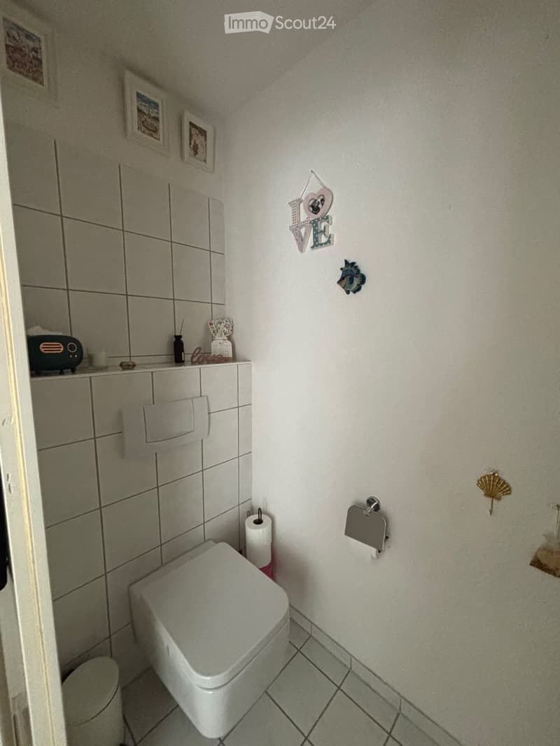 A small bathroom with a toilet, toilet paper holder, and decorative wall art including a 'LOVE' sign and framed pictures. The walls are tiled, and there is a radiator on the left side.