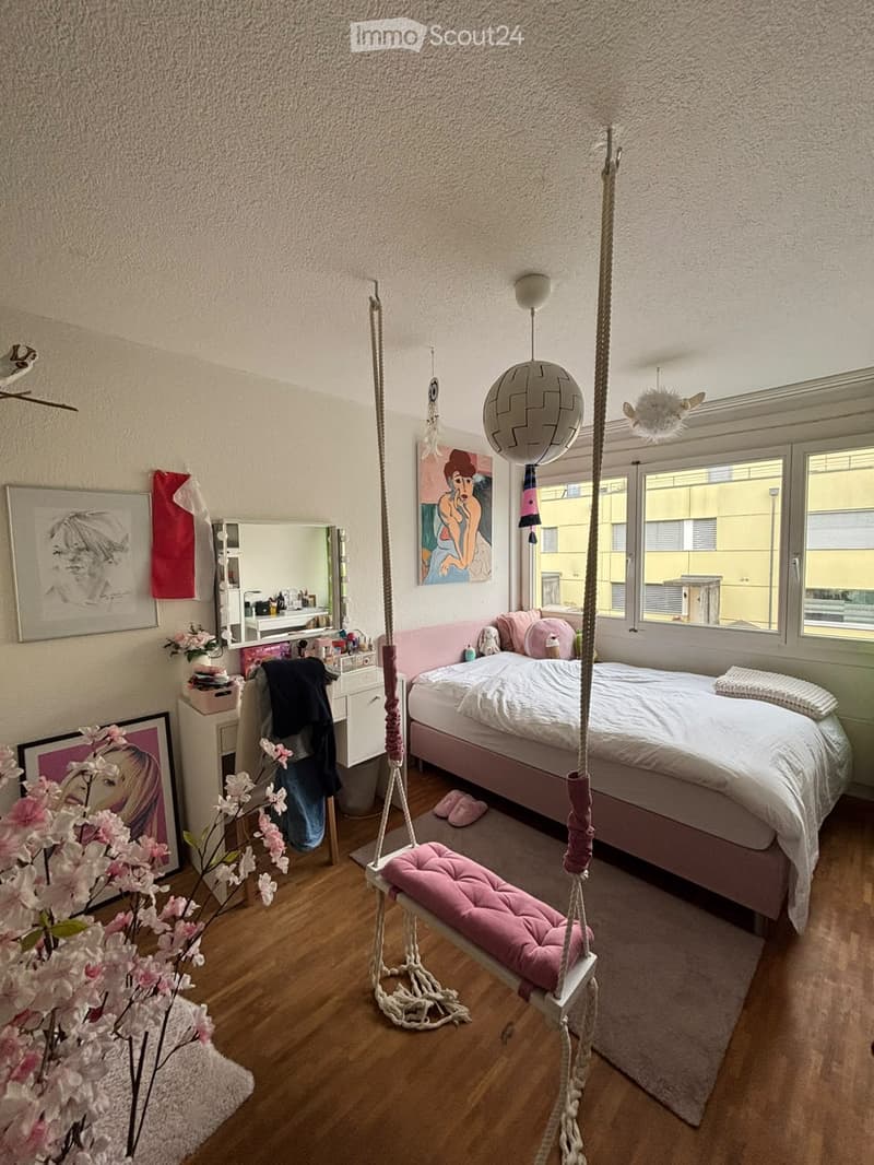Cozy bedroom with a pink bed, white bedding, a wicker hanging chair, framed artwork, a mirror, and a flower arrangement on the floor.