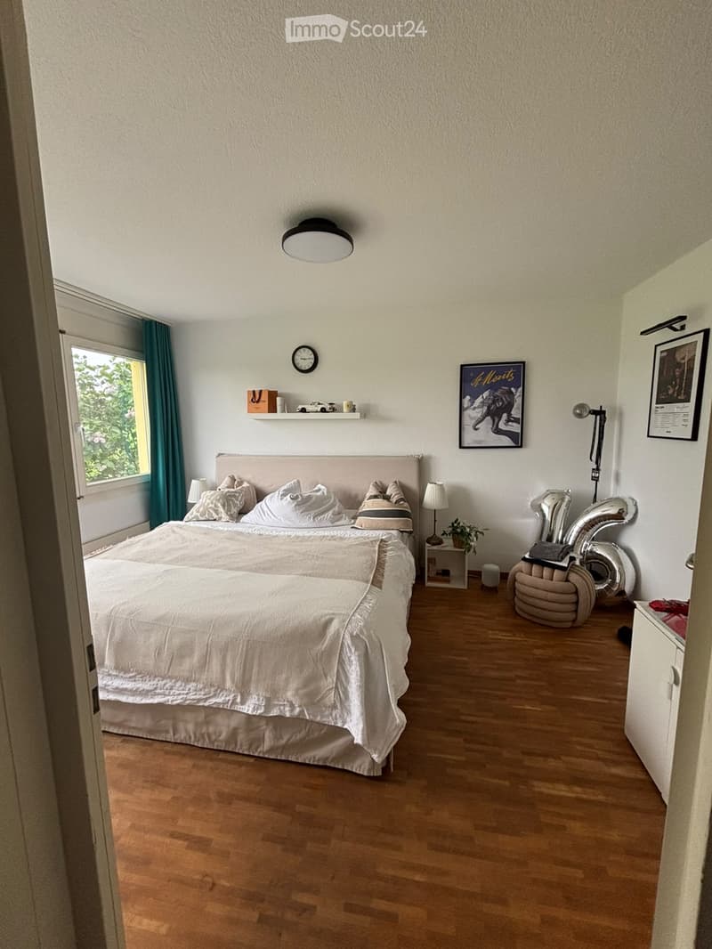 Well-lit bedroom with a large bed, nightstand, lamp, picture frames, and a shelf with decorative items.