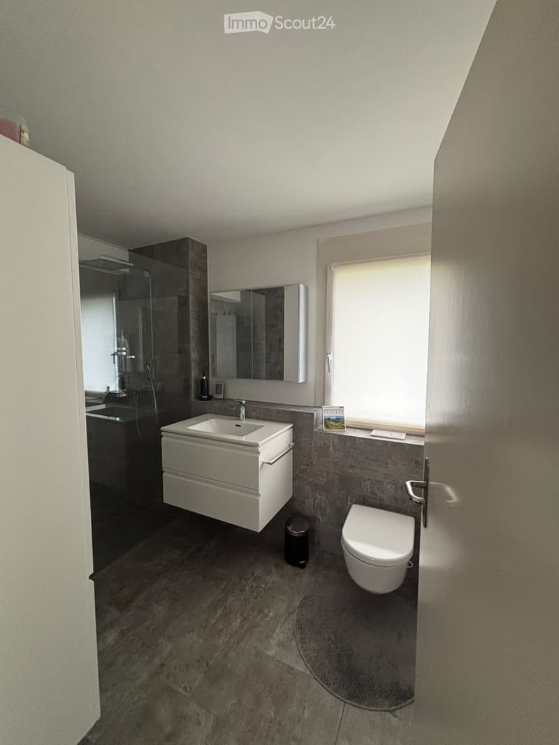 Modern bathroom with a white sink, toilet, mirror, and a large window with blinds. The floor is tiled in a dark grey marble pattern.