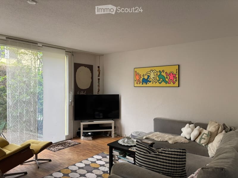 Modern living room with a grey sectional sofa, coffee table, yellow armchair, television on a stand, and artwork on the walls. The room features large windows with blinds and a view of greenery outside.