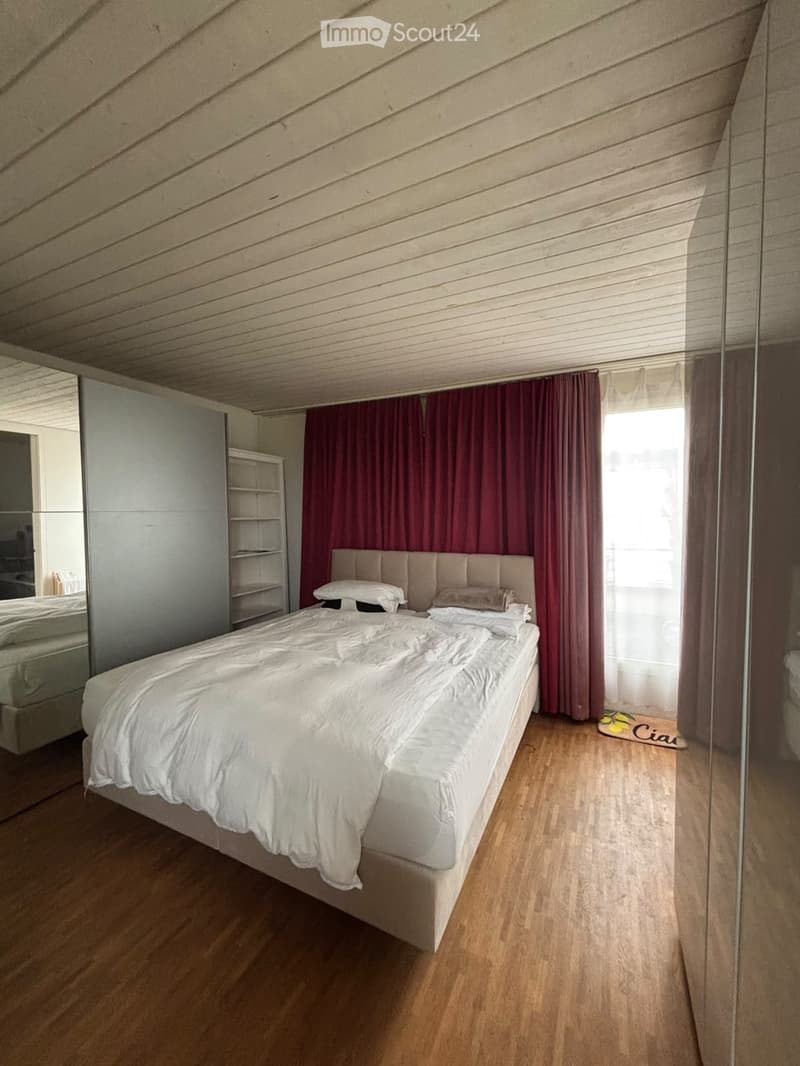 Modern bedroom with a large bed, headboard, pillows, a mirrored wardrobe, and a shelf. The room features wooden flooring and red curtains.