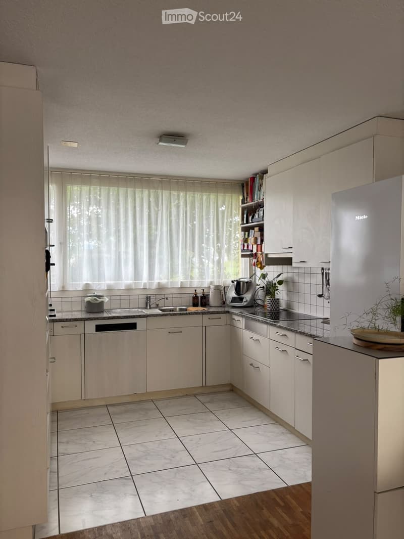 Modern kitchen with light-colored cabinets, marble tile floor, and wooden countertop. Features a refrigerator, sink, blender, and various kitchen items.