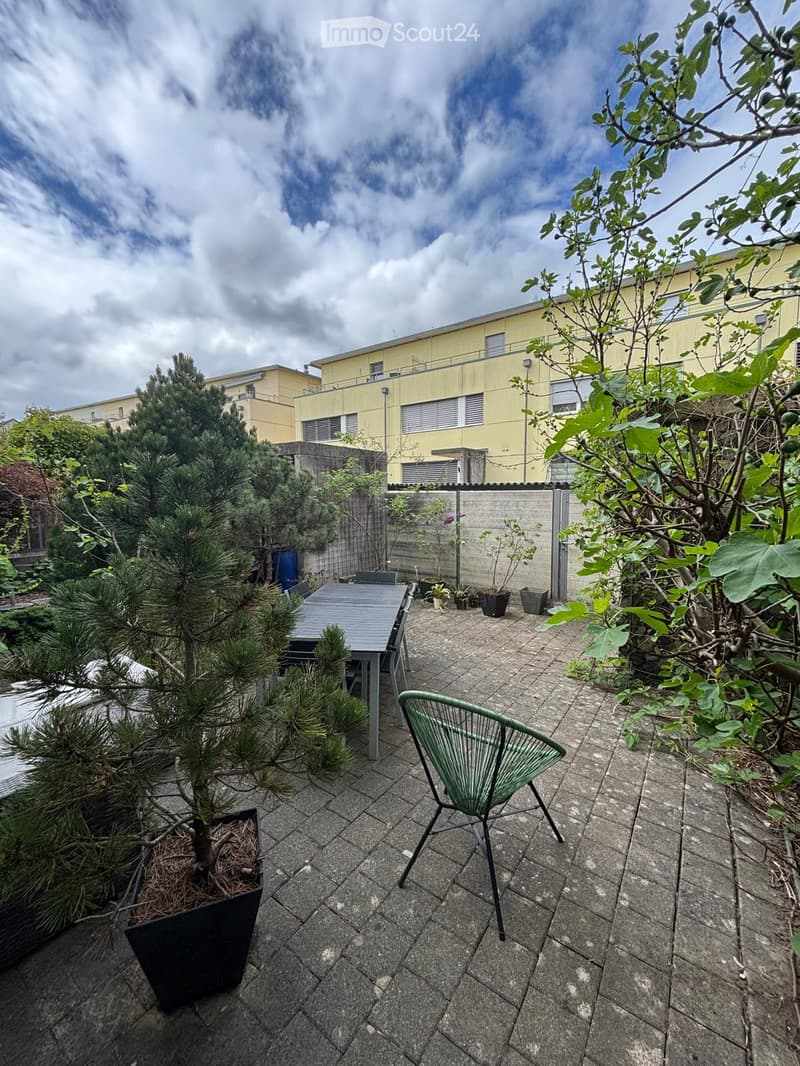 A small, paved outdoor area with a table, chair, and several potted plants, surrounded by trees and a building in the background.