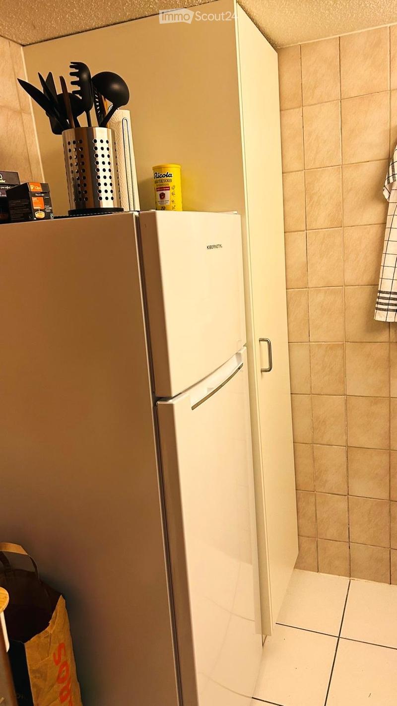 A small kitchen with a white refrigerator, beige tiled walls, and a utensil holder on top of the fridge.