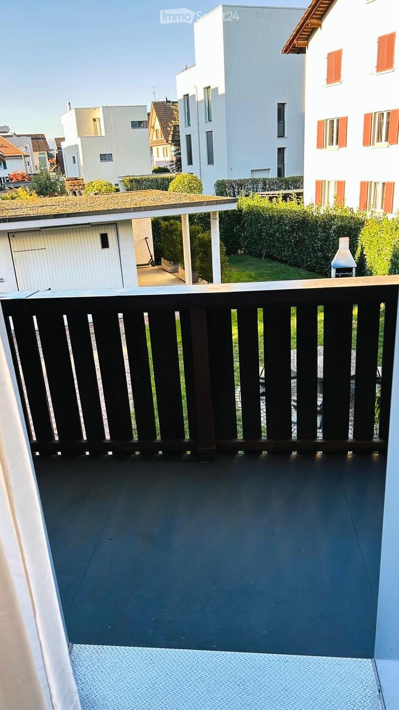 Balcony with dark wooden railing, grey tiled floor, and a view of a residential area with houses, greenery, and a garage.