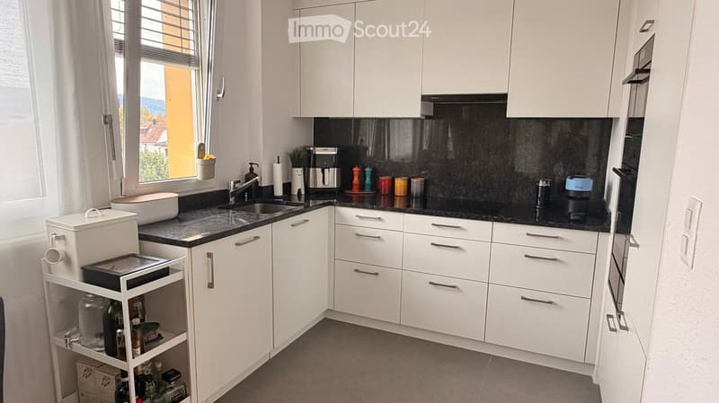 Modern kitchen with white cabinets, black countertops, and various appliances including a coffee maker, sink, and bottle rack. The space features a window with blinds and a small potted plant on the windowsill.