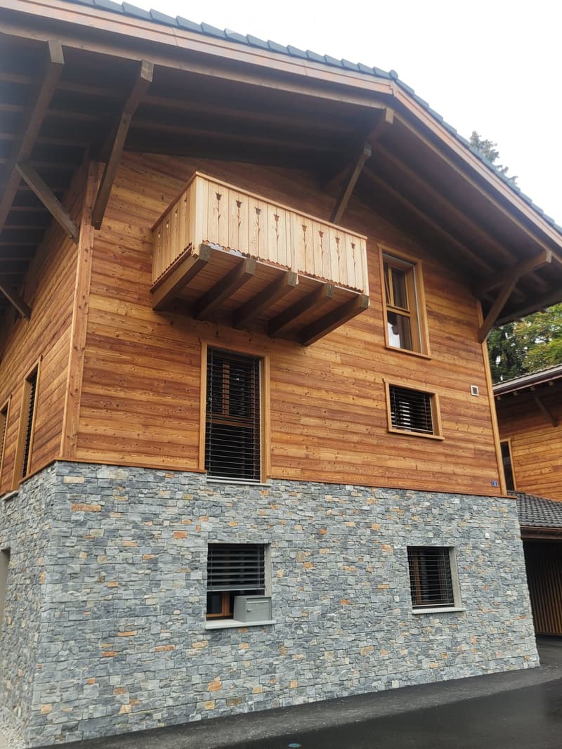 A two-story house with a combination of wooden and stone exterior walls, featuring large windows, a balcony, and a raised ground floor.