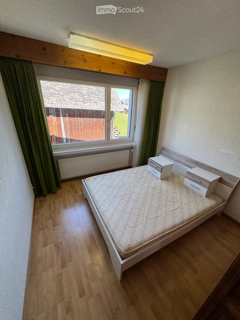 A tidy bedroom with a single bed, nightstand, and drawer, featuring wooden flooring and large windows with green curtains.