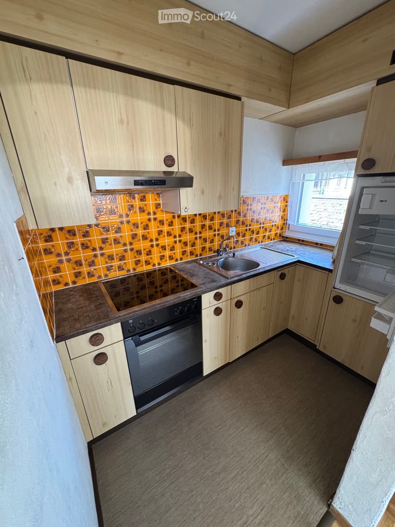 Modern kitchen with light wooden cabinets, stainless steel sink, oven, and refrigerator. Features a tiled backsplash and a window providing natural light.