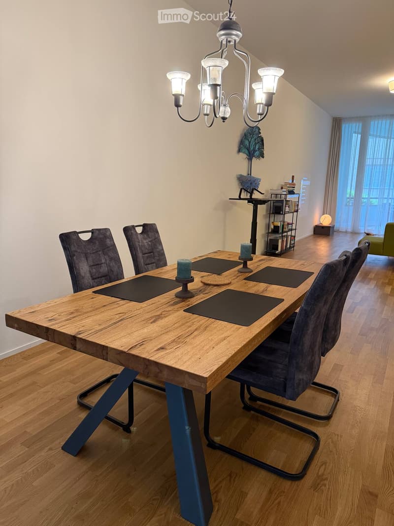 Dining room with a wooden table, four chairs, and a chandelier hanging from the ceiling. There are candles on the table and a shelf with books in the background.
