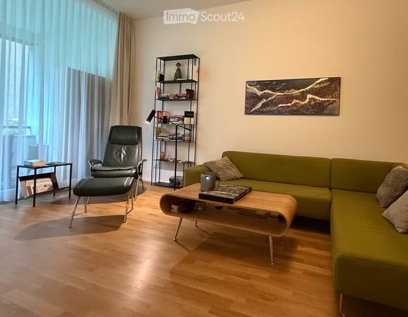 Modern living room with a green sectional sofa, wooden coffee table, black leather chair, bookshelf, and large windows with curtains.