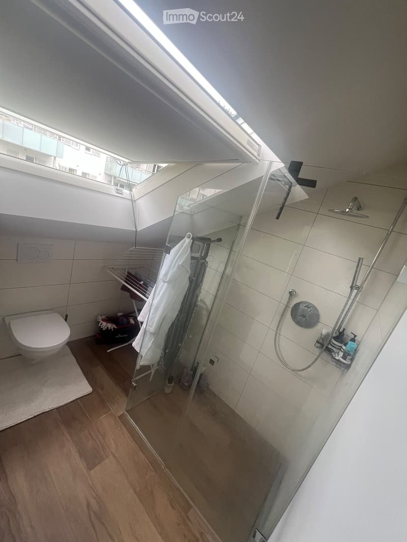 Modern bathroom with a toilet, glass-enclosed shower, towel rack, and shower rack. The room features wooden flooring and white tiled walls.