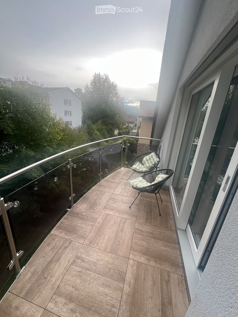 Balcony with wooden floor, glass railing, and two chairs with green cushions.