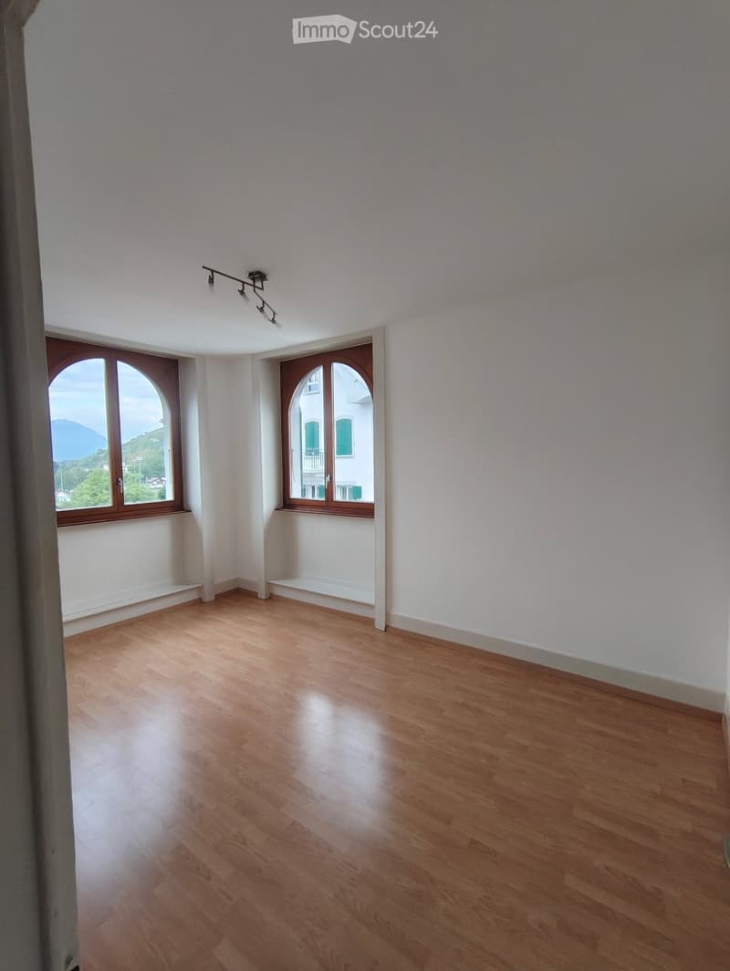 Empty room with light wooden flooring, white walls, and two arched windows offering a scenic view.