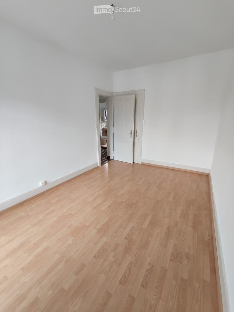 Empty room with light wooden flooring and white walls, featuring an open doorway leading to another room.