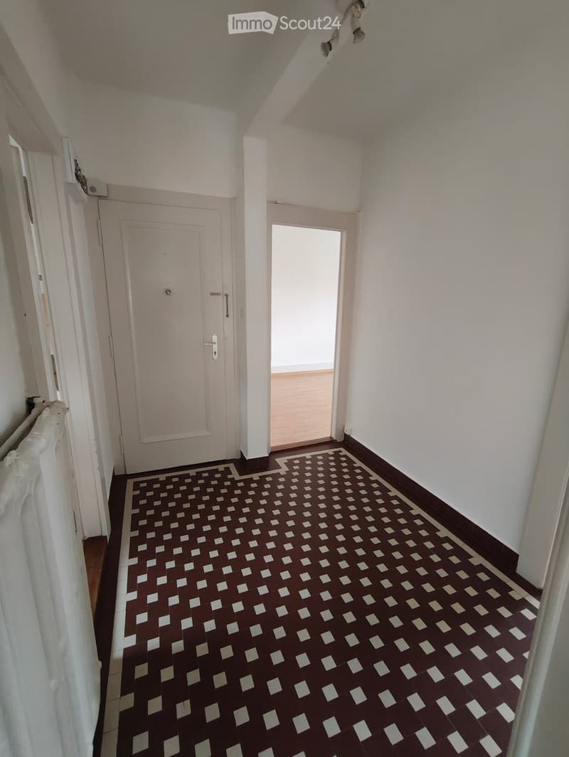 A narrow hallway with patterned tiles, white walls, and a closed door on the left and an open doorway on the right.