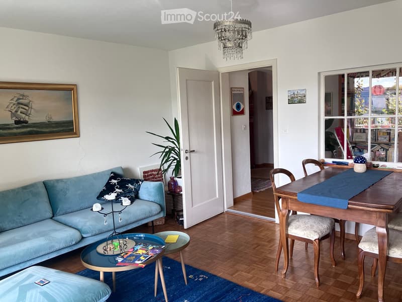 Bright living room with a blue sofa, coffee table, dining table, chairs, and a chandelier. Features a painting, plant, and decorative items.