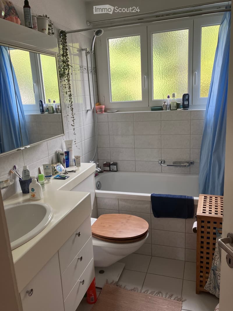 Bathroom with a sink, toilet, bathtub, and mirror. The sink has various toiletries, and there is a towel on the edge of the bathtub. The room features tiled walls and a window with frosted glass.