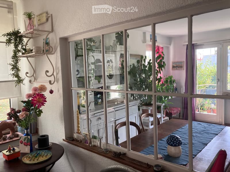A cozy, well-lit room with a dining area and an open kitchen. The dining area features a wooden table with chairs, a blue rug, and decorative items including a mushroom-shaped stool. The kitchen has white cabinets, a sink, and various decorative plants vi